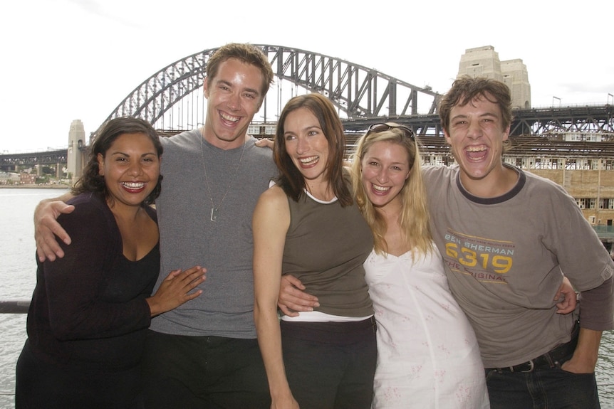 Three women and two men pose hugging each other while laughing and smiling with the Sydney Harbor Bridge in the background